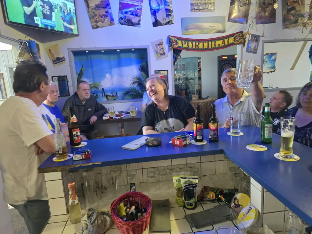Sechs Leute sitzen und stehen um eine Bar herum, trinken und unterhalten sich, im Hintergrund sind Plakate und Flaschen zu sehen.