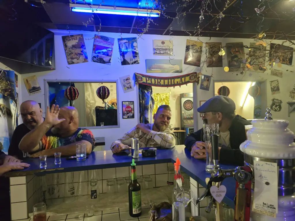 Vier Männer unterhalten sich und lachen an einer mit Plakaten und einem „Portugal“-Banner dekorierten Bar, Getränke stehen auf der Theke.