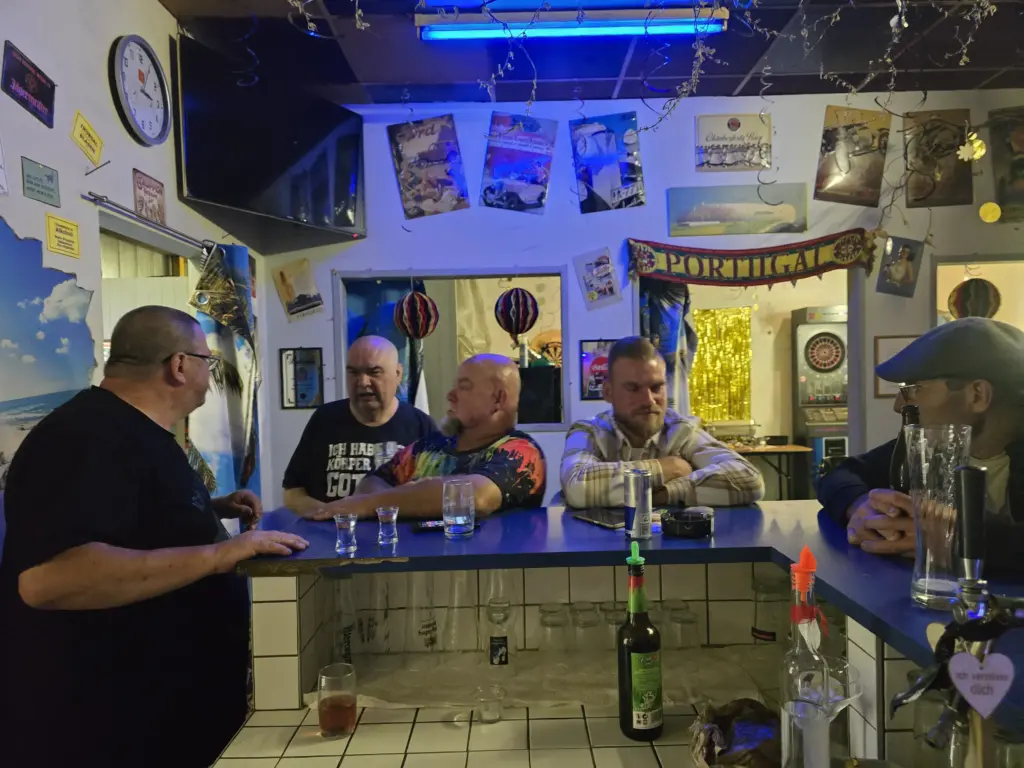 Fünf Männer unterhalten sich in einer Bar, die mit Reiseplakaten, Lichtern und einem „Portugal“-Banner dekoriert ist.