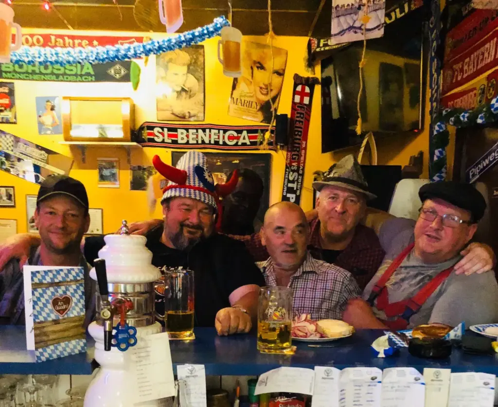 Fünf Männer posieren und lächeln an einer Bar, die mit Fußballschals und Bier, festlichen Hüten und bayerischem Dekor dekoriert ist.