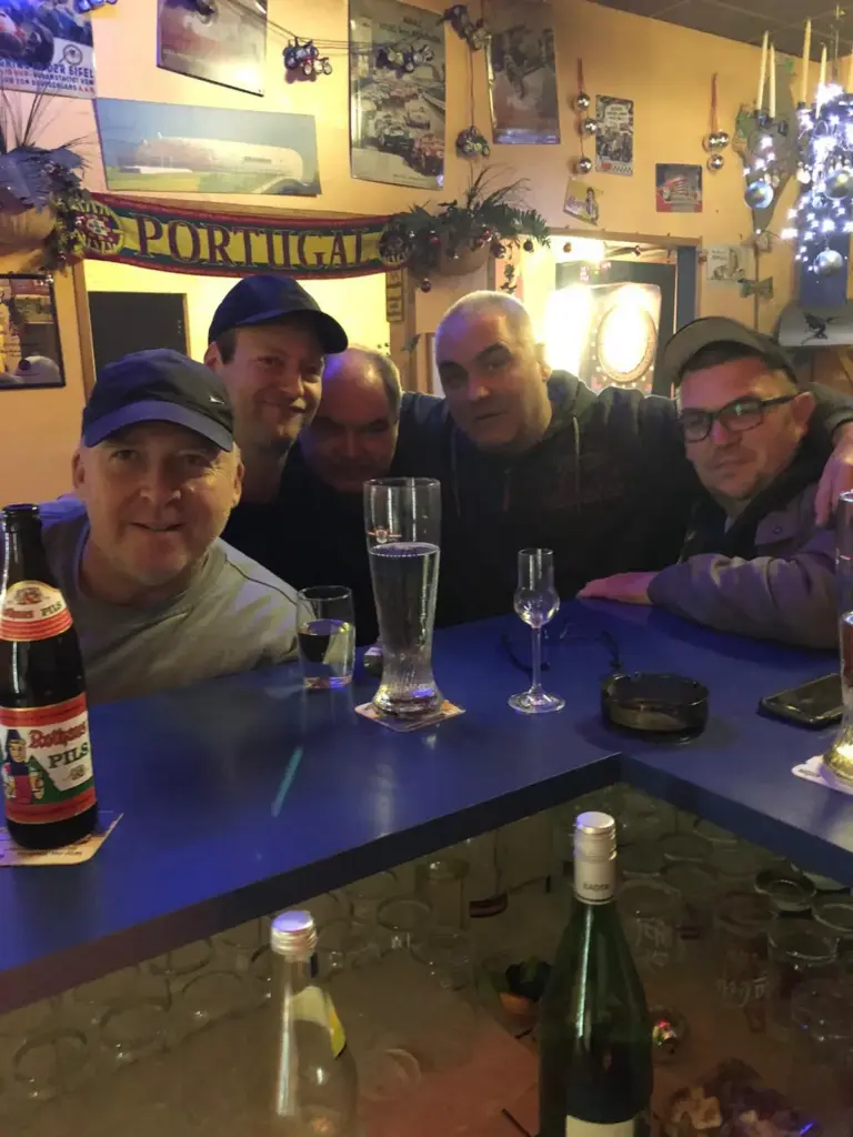 Fünf Männer sitzen lächelnd mit Getränken an einer Bar in einem mit Plakaten und einer „PORTUGAL“-Flagge dekorierten Pub.