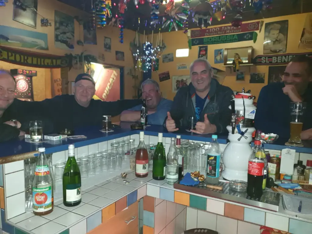 Fünf Männer sitzen lächelnd und trinkend an einer farbenfrohen Bar, umgeben von Flaschen und festlicher Dekoration.