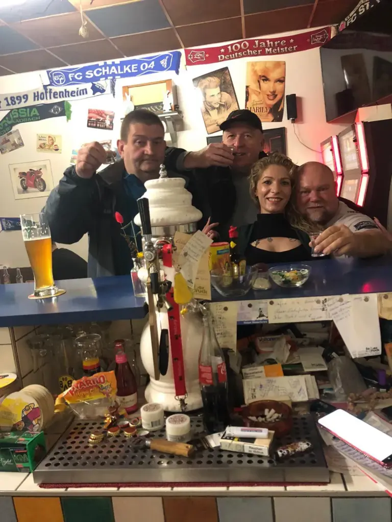 Vier Personen posieren und lächeln gemeinsam hinter einer überfüllten Bartheke mit Getränken und Snacks.