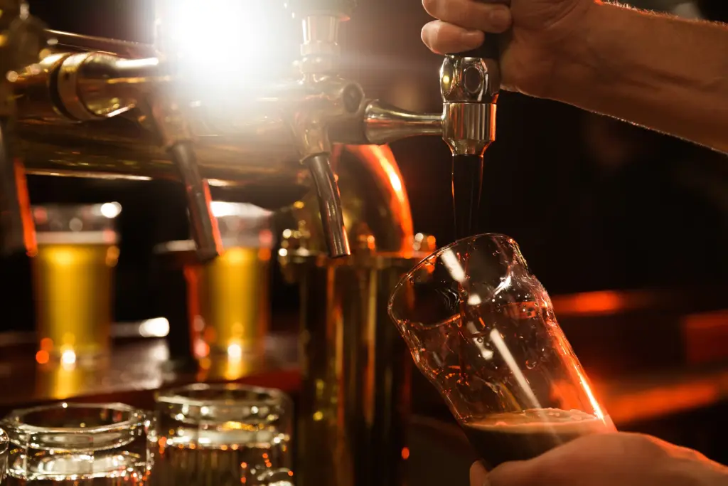 An einer Bar wird dunkles Bier von Hand vom Zapfhahn in ein Glas gezapft, im Hintergrund sind andere Getränke zu sehen – ein perfekter Moment für jeden Getränkehandel in March-Hugstetten.